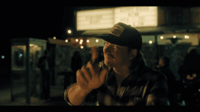 A Man In A Baseball Cap Stands In Front Of A Sign That Says ' Live Free ' GIF