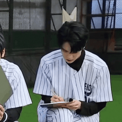 A Man In A Baseball Uniform Is Sitting On The Grass Writing On A Clipboard . GIF