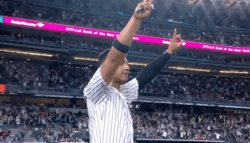 A Man In A Baseball Uniform Stands In Front Of An Official Bank Of The New York Sign GIF