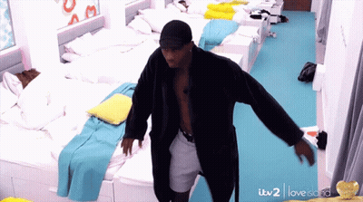 A Man In A Bathrobe Is Dancing In A Room With Lots Of Beds And A Sign That Says Love Island GIF