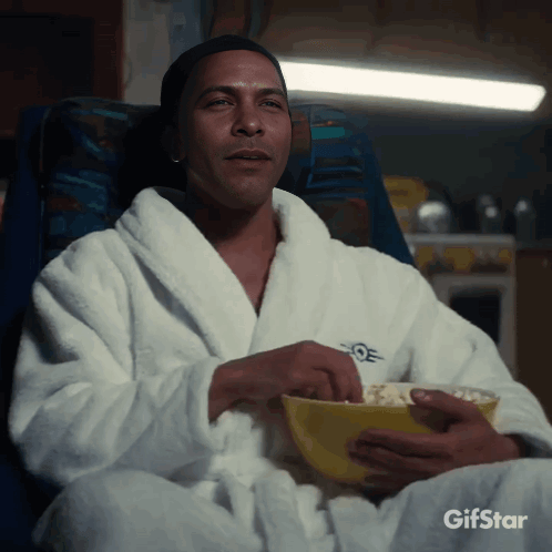 A Man In A Bathrobe Is Eating Popcorn From A Bowl GIF
