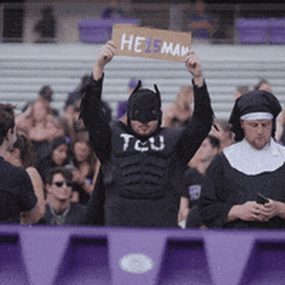 A Man In A Batman Costume Is Holding Up A Sign That Says Heisman GIF