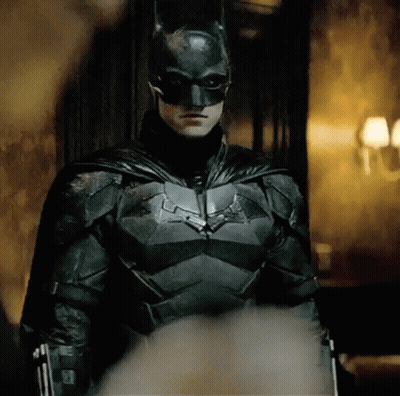A Man In A Batman Costume Is Standing In A Room With A Gun . GIF