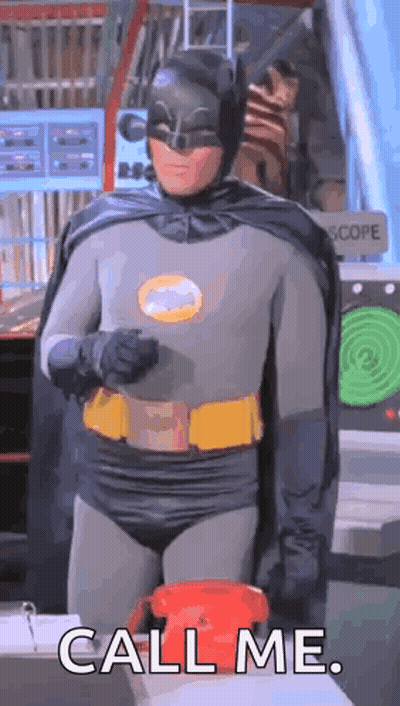 A Man In A Batman Costume Is Standing In Front Of A Red Telephone And Saying `` Call Me '' . GIF