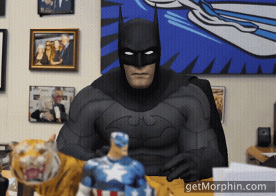 A Man In A Batman Costume Sits At A Desk With A Tiger And A Captain America Figurine GIF