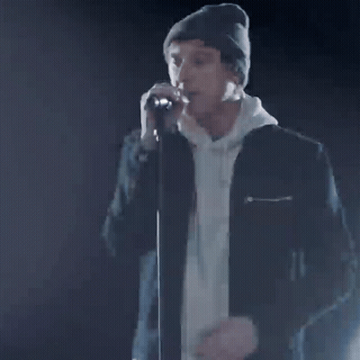 A Man In A Beanie Is Singing Into A Microphone With The Words Our Time Written On It . GIF