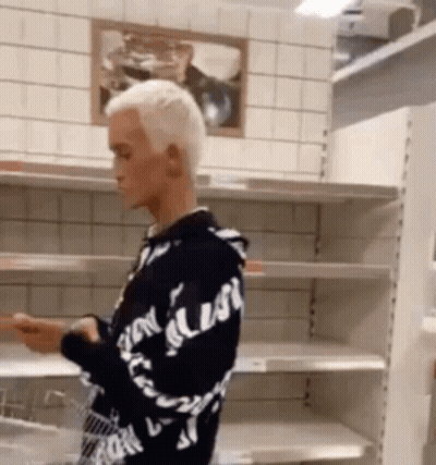A Man In A Black And White Hoodie Is Standing In Front Of Empty Shelves . GIF