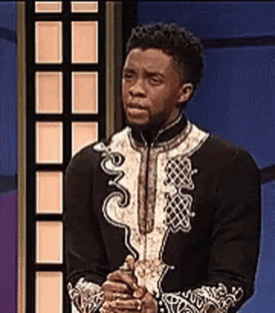 A Man In A Black And White Outfit Is Standing In Front Of A Blue Wall With His Hands Folded . GIF