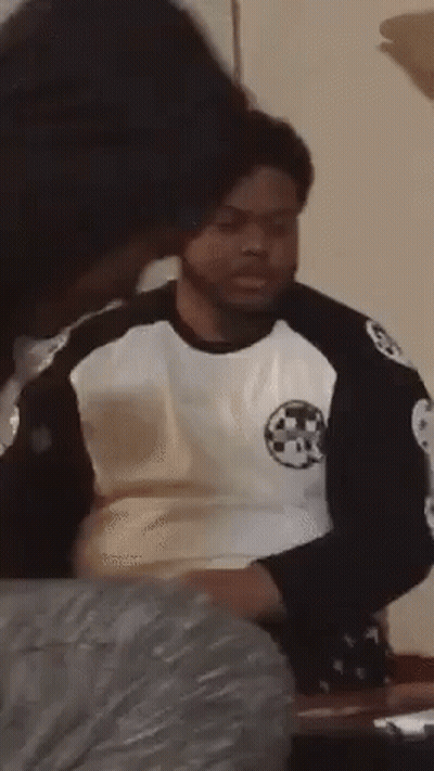 A Man In A Black And White Shirt Is Sitting At A Table With A Woman Behind Him . GIF
