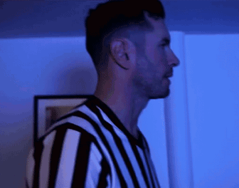 A Man In A Black And White Striped Shirt Stands In A Dark Room GIF