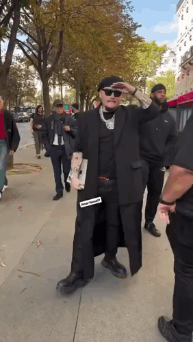A Man In A Black Coat And Hat Is Walking Down A Sidewalk With A Group Of People . GIF