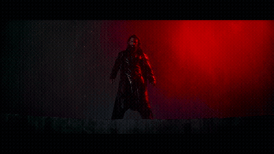 A Man In A Black Coat Is Standing In A Dark Room With Red Lights Behind Him . GIF