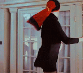 A Man In A Black Coat Is Standing In Front Of A French Door Holding A Red Pillow On His Head . GIF