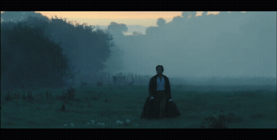 A Man In A Black Coat Stands In A Field With Trees In The Background GIF