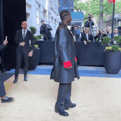 A Man In A Black Coat With Red Cuffs Stands In Front Of A Crowd GIF