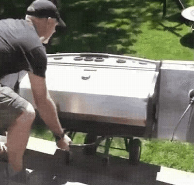 A Man In A Black Hat Is Pushing A Cart With A Grill In The Background GIF