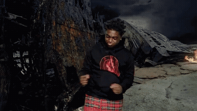 A Man In A Black Hoodie And Plaid Shorts Is Standing In Front Of A Burned Out Building . GIF