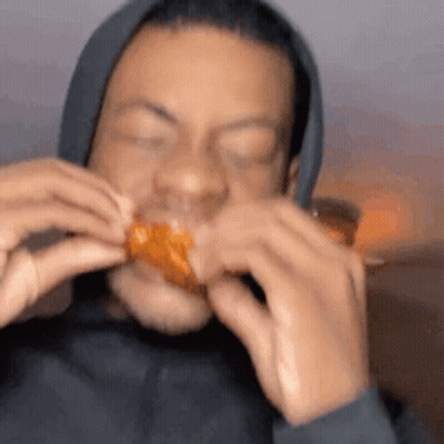 A Man In A Black Hoodie Is Eating A Chicken Wing With His Eyes Closed . GIF