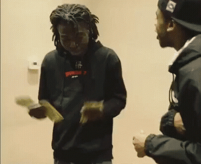 A Man In A Black Hoodie Is Holding A Bunch Of Money While Standing Next To Another Man In A Hat . GIF