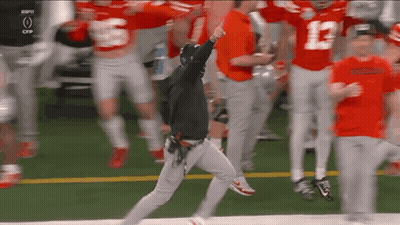 A Man In A Black Hoodie Is Running On A Football Field Surrounded By Football Players . GIF