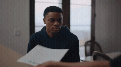 A Man In A Black Hoodie Is Sitting At A Table With A Piece Of Paper In Front Of Him GIF