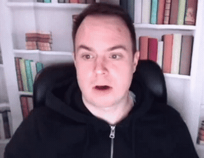A Man In A Black Hoodie Is Sitting In Front Of A Bookshelf And Making A Funny Face . GIF