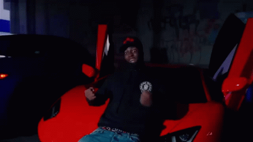 A Man In A Black Hoodie Is Sitting In Front Of A Red Sports Car . GIF