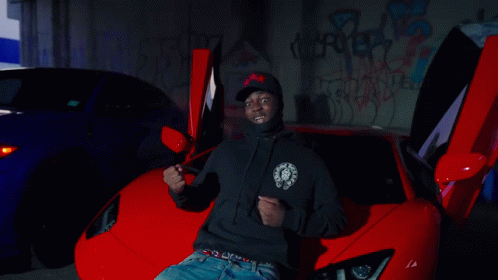 A Man In A Black Hoodie Is Sitting In Front Of A Red Sports Car GIF