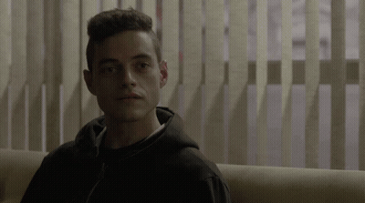 A Man In A Black Hoodie Is Sitting On A Couch In Front Of Vertical Blinds GIF