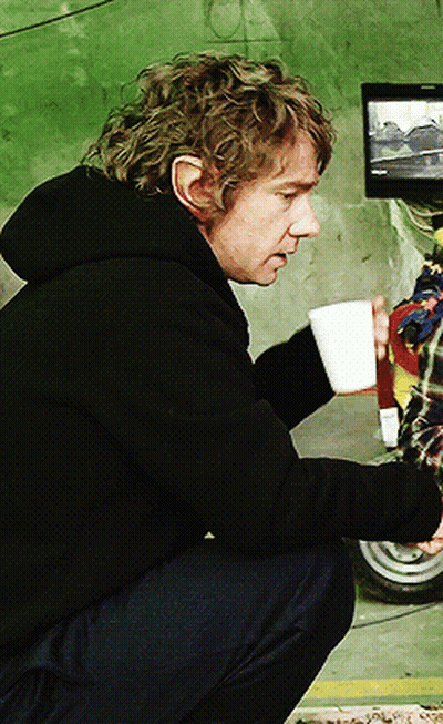 A Man In A Black Hoodie Is Squatting Down Holding A Cup Of Coffee GIF
