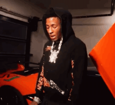 A Man In A Black Hoodie Is Standing In Front Of An Orange Car In A Garage . GIF