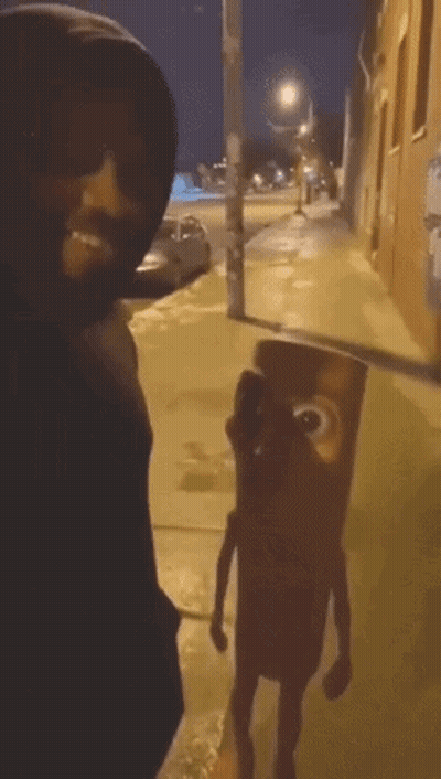 A Man In A Black Hoodie Is Standing Next To A Cartoon Character On The Sidewalk GIF