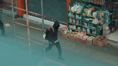 A Man In A Black Hoodie Is Walking Down A Sidewalk In Front Of A Store . GIF