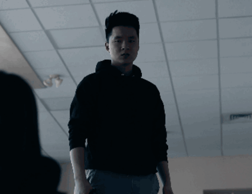 A Man In A Black Hoodie Stands In A Dark Room With The Word Sure Behind Him GIF