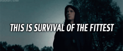 A Man In A Black Hoodie Stands In Front Of Trees With The Words " This Is Survival Of The Fittest " GIF