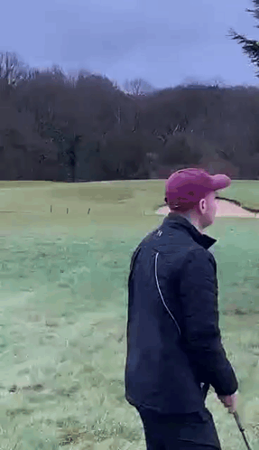 A Man In A Black Jacket And A Red Hat Is Walking On A Golf Course Holding A Golf Club . GIF