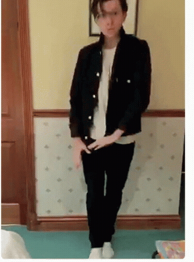A Man In A Black Jacket And Black Pants Is Standing In Front Of A Wall . GIF