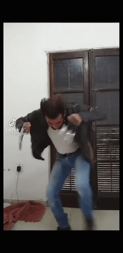 A Man In A Black Jacket And Blue Jeans Is Jumping In The Air While Holding A Gun . GIF