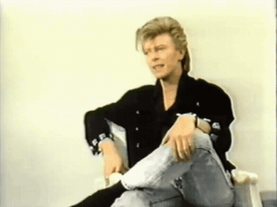 A Man In A Black Jacket And Jeans Is Sitting In A Chair With His Legs Crossed . GIF