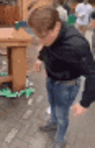 A Man In A Black Jacket And Jeans Is Standing On A Sidewalk . GIF