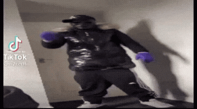 A Man In A Black Jacket And Purple Gloves Is Standing In A Room . GIF