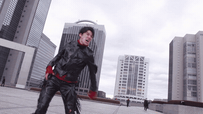 A Man In A Black Jacket And Red Gloves Stands In Front Of A City Skyline GIF