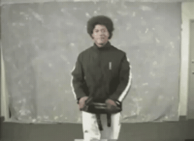 A Man In A Black Jacket And White Pants Is Standing In Front Of A White Wall Holding A Black Belt . GIF