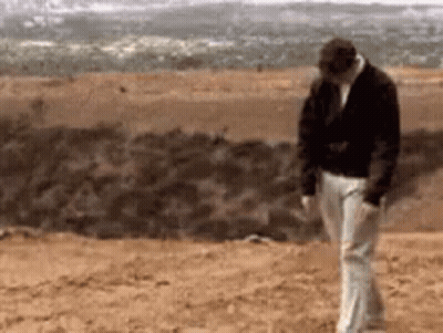 A Man In A Black Jacket And White Pants Is Walking On A Dirt Field . GIF