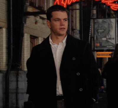 A Man In A Black Jacket And White Shirt Is Walking Down A Street In Front Of A Sign That Says ' Coca Cola ' GIF