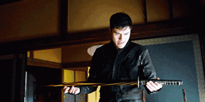 A Man In A Black Jacket Holds A Large Sword In His Hands GIF