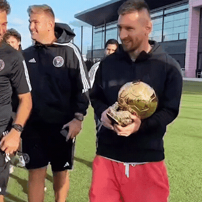 A Man In A Black Jacket Is Holding A Gold Soccer Ball While Standing On A Field . GIF