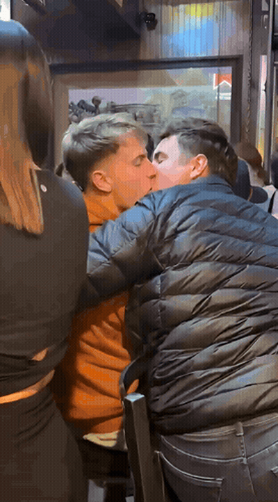 A Man In A Black Jacket Is Kissing Another Man In An Orange Shirt GIF