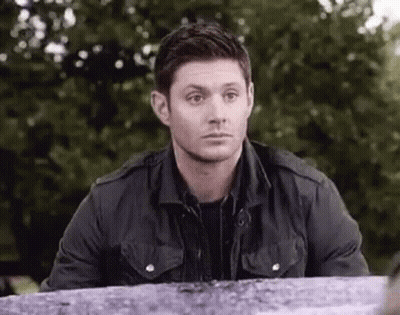 A Man In A Black Jacket Is Sitting At A Table In A Park And Looking At The Camera . GIF