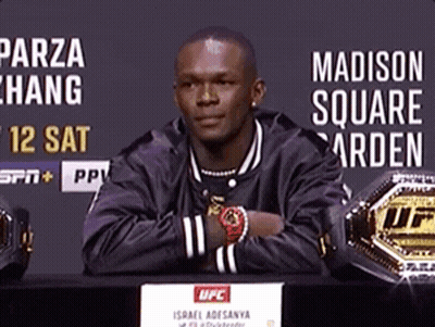 A Man In A Black Jacket Is Sitting At A Table With His Arms Crossed In Front Of A Sign That Says Madison Square Garden GIF